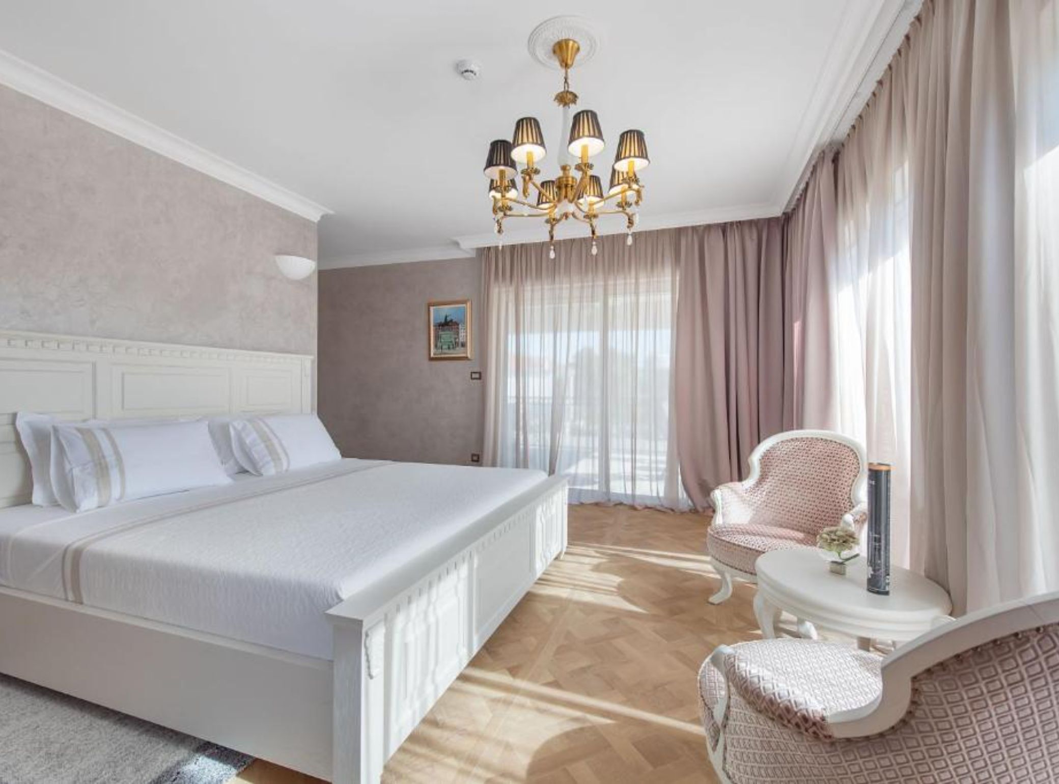 A bedroom with a large bed, a small side table, a chair, a chandelier, and a window with curtains.