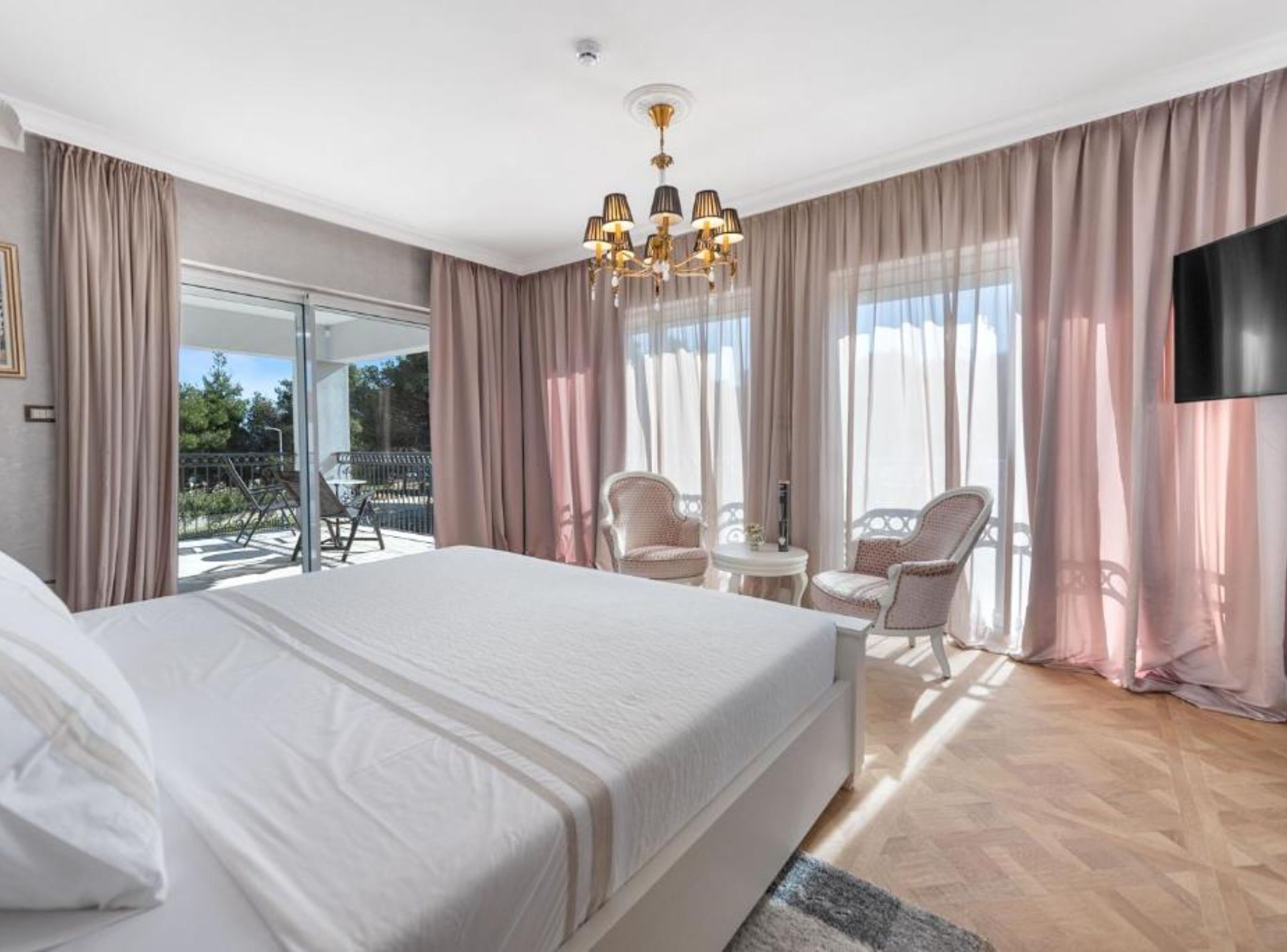 Habitación de hotel con una cama grande, balcón con sillas, candelabro y televisor en la pared.