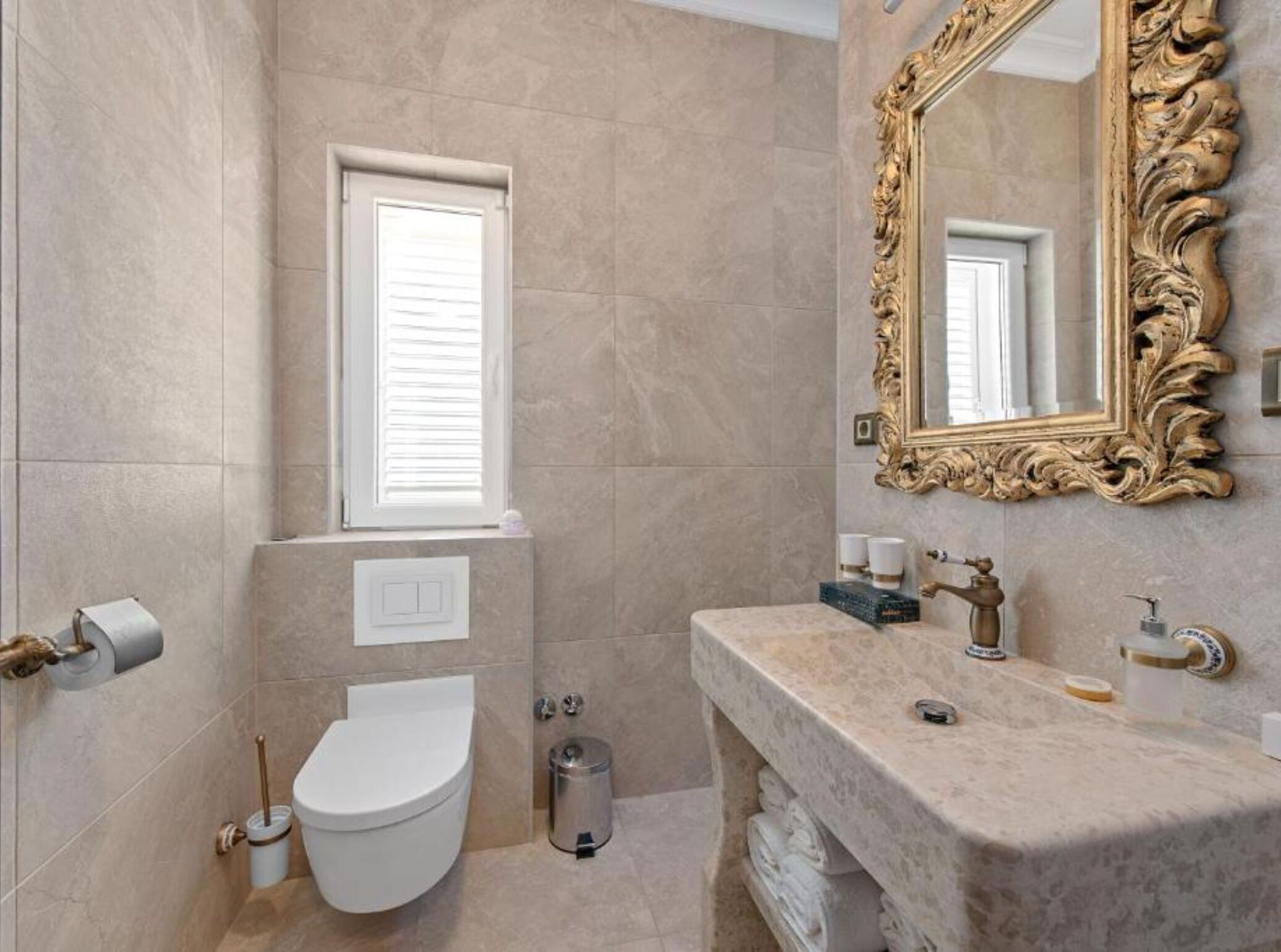 Ein Badezimmer mit beigen Fliesen, einer weißen Toilette, einem Waschbecken mit Marmorplatte, einem spiegel mit goldener Rahmung und einem Fenster mit Jalousien.
