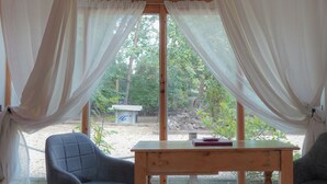 Ein Zimmer mit Holztisch und zwei Stühlen, einem Fenster mit transparenten Vorhängen und Blick auf einen Garten.