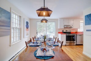 Un comedor con una mesa de madera dispuesta para seis personas, una lámpara colgante y una cocina con electrodomésticos de acero inoxidable.