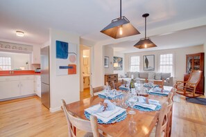 A dining area with a wooden table set for six, a kitchen with white cabinets, and a living room with a sofa and a large window.