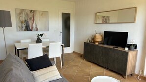 Un salon comprenant un canapé, une table basse, un téléviseur posé sur un meuble en bois, une table à manger avec des chaises, et des œuvres d’art accrochées aux murs.