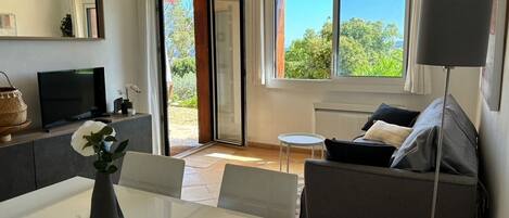 A modern living room with a sofa, a dining table with a vase, a TV, and a large window with a view of greenery.