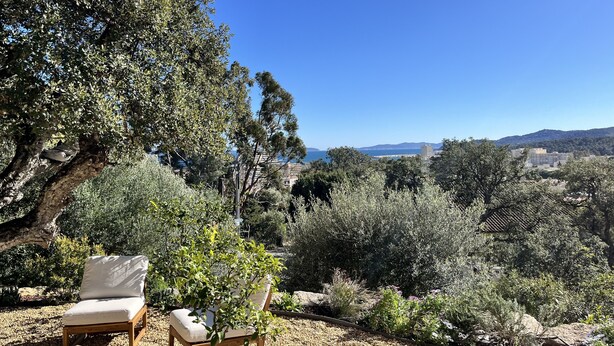Une terrasse en bois avec deux chaises longues et une table, offrant une vue panoramique sur un paysage verdoyant, avec des arbres et une vue lointaine sur la mer.