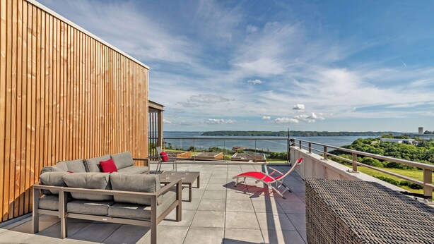 A terrace with a sofa, chairs, and a view of a lake and distant buildings. - Rivages Appartement Pleine vue mer Terrasse Privative et Garage (Brest)
