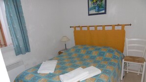 A bed with a patterned bedspread, a white chair, a nightstand with a lamp, and a painting on the wall.