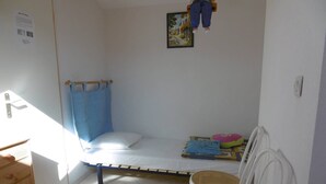A small, bright room with a single bed, two chairs, a small table, and a window with sunlight.