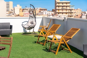 Een terras op het dak met kunstgras, houten lounge stoelen, een hangstoel en een grijze bank.