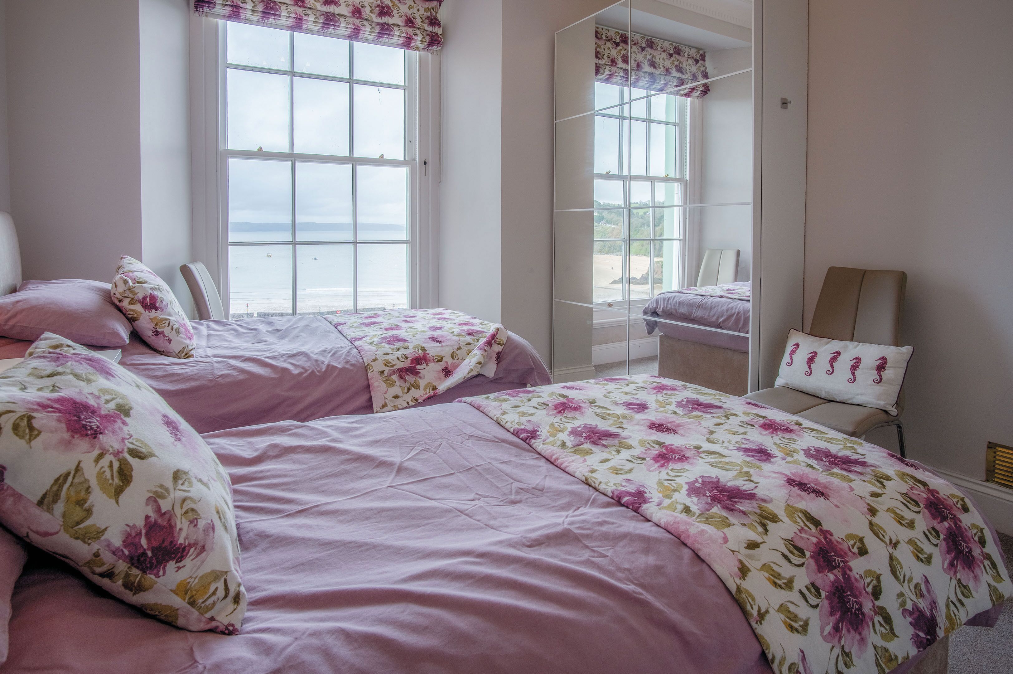 Une chambre avec deux lits, une literie à motifs floraux, une grande fenêtre et une vue sur la mer.