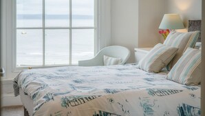 A bedroom with a large window overlooking the sea, a bed with striped bedding, a chair, and a bedside table with a lamp and flowers.