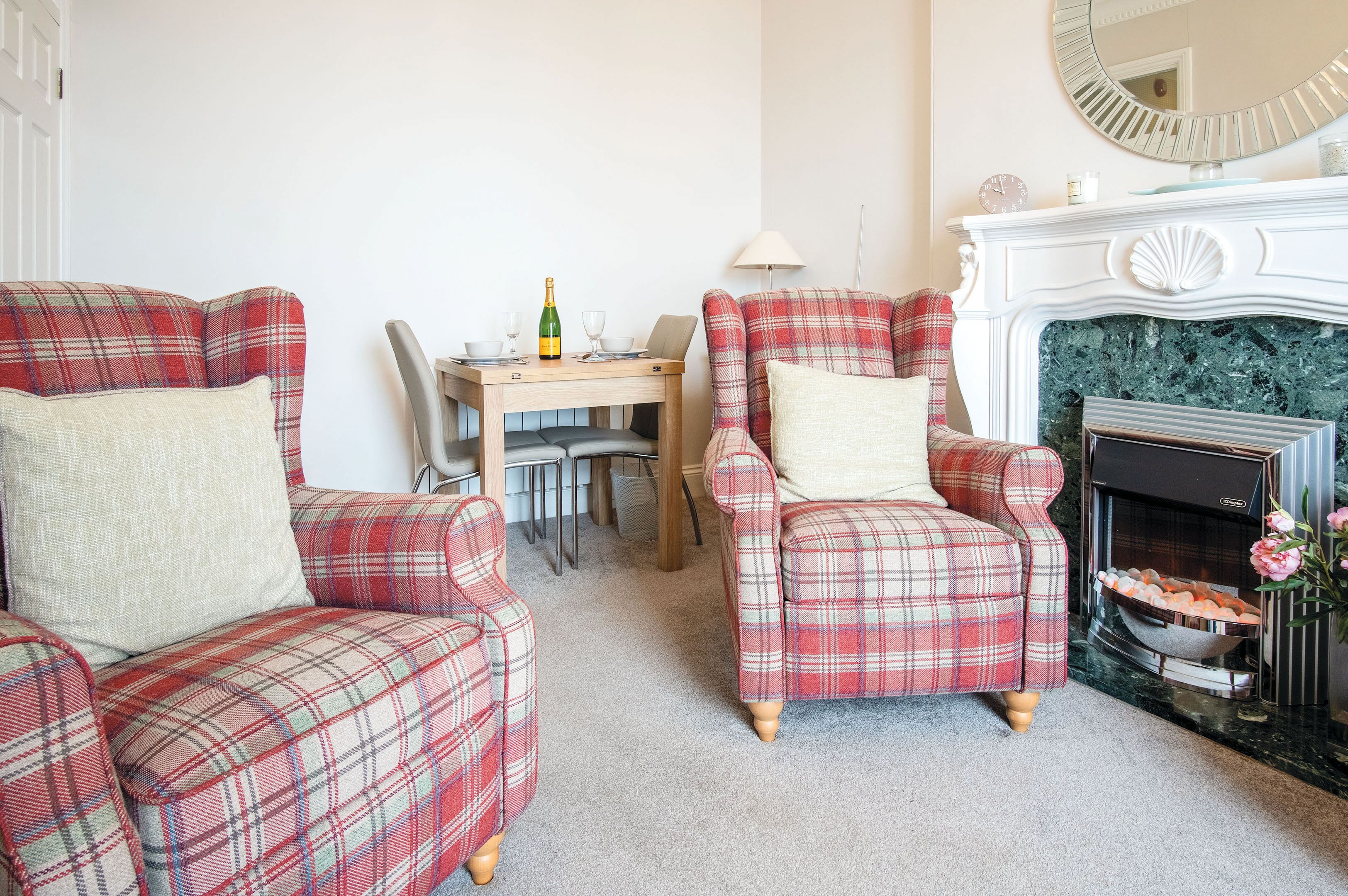 A living room with two plaid armchairs, a small table with a bottle and glasses, a fireplace, and a mirror on the wall.