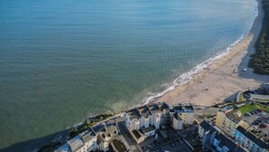An aerial view of a coastal town with a sandy beach, a clear blue sea, and buildings along the shore.