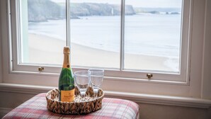A bottle of champagne and two glasses on a woven tray on a plaid cushion by a window with a view of a beach and cliffs.