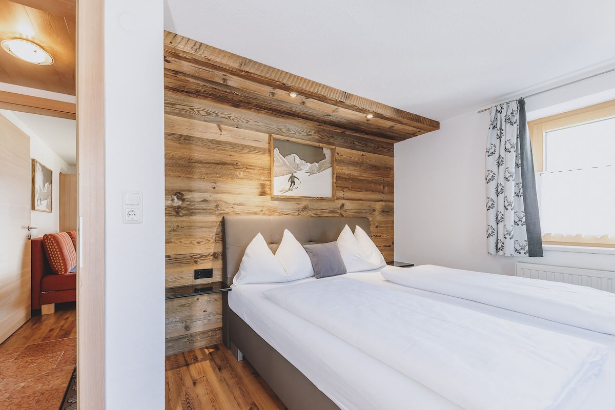 A bedroom with a wooden headboard, a bed with white bedding, a bedside table, and a window with curtains.