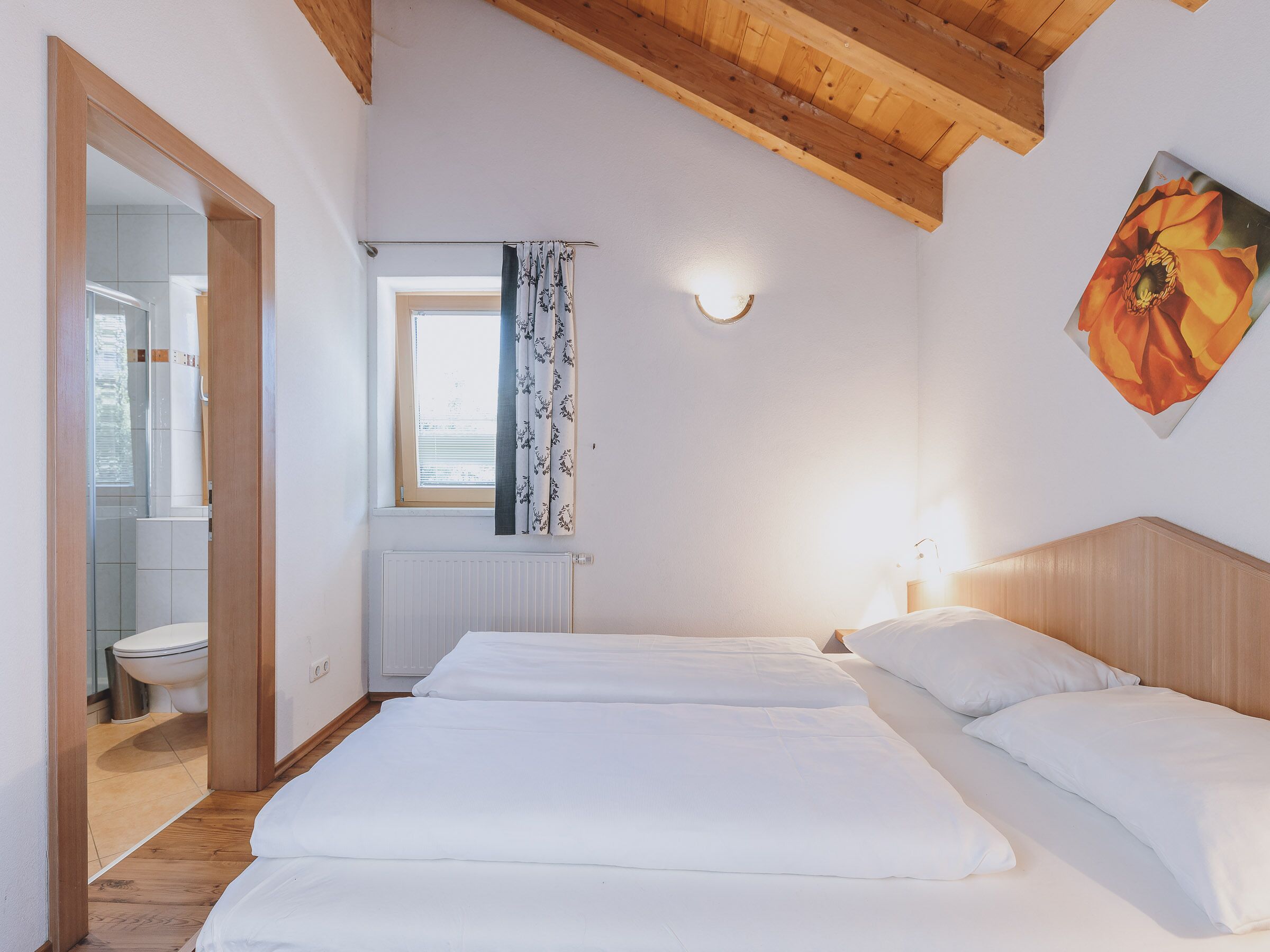 Een slaapkamer met twee bedden, een raam met gordijnen, een radiator en een houten plafond.