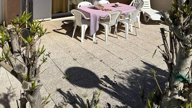 Eine Terrasse mit Tisch und Stühlen, ein überdachter Bereich und ein Gebäude mit einem Ziegeldach und verstellbaren Fensterläden.