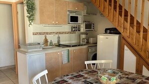 Eine kleine Küche mit Holzausstattung, einer Mikrowelle, einem Toaster und einem Kühlschrank. Ein Esstisch mit Stühlen und einer dekorativen Tischläufer ist ebenfalls vorhanden. Im Hintergrund ist eine Holztreppe zu sehen.