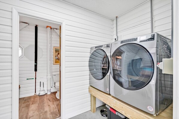 Une buanderie équipée de deux machines à laver à chargement frontal, posées sur une plateforme en bois, avec une fenêtre et une porte donnant sur une salle de bain.