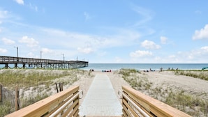 A wooden boardwalk leading to a beach with a pier in the background. - Spacious 5BR, 4BA home at Holden Beach with stunning water views. (Holden Beach)