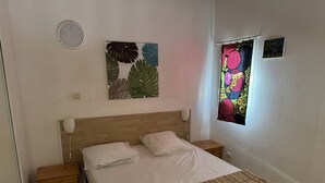 Ein Schlafzimmer mit einem Holz-Kopfteil, zwei Nachttischen, einem Bett mit weißer Bettwäsche, einer gemusterten Tagesdecke, einer Wandlampe und einem Fenster mit einem farbenfrohen Vorhang.