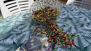 Ein Tisch mit Tischdecke mit tropischem Blattmuster, eine dekorative Glasschale mit farbigem Folieninhalt und eine Karte mit dem Schriftzug 'HOLIDAYLAND'.