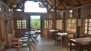 Un comedor rústico de madera con mesas y sillas, ventanas amplias con vistas al verdor, y un techo alto con vigas a la vista.