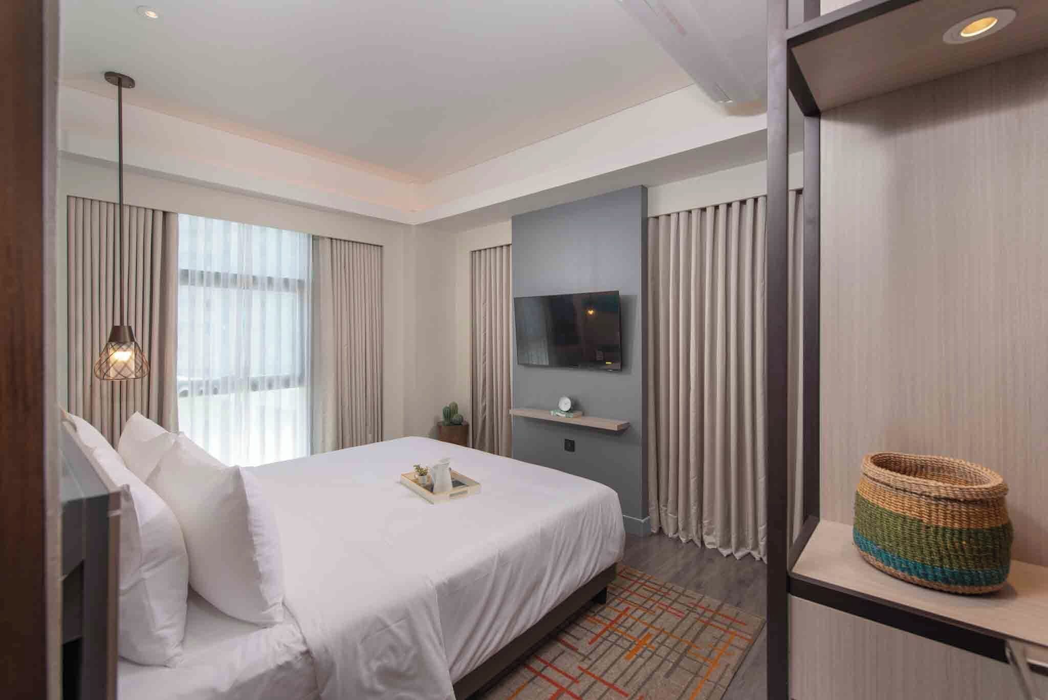 A hotel room with a bed, a television, a small table, a plant, and a window with curtains.