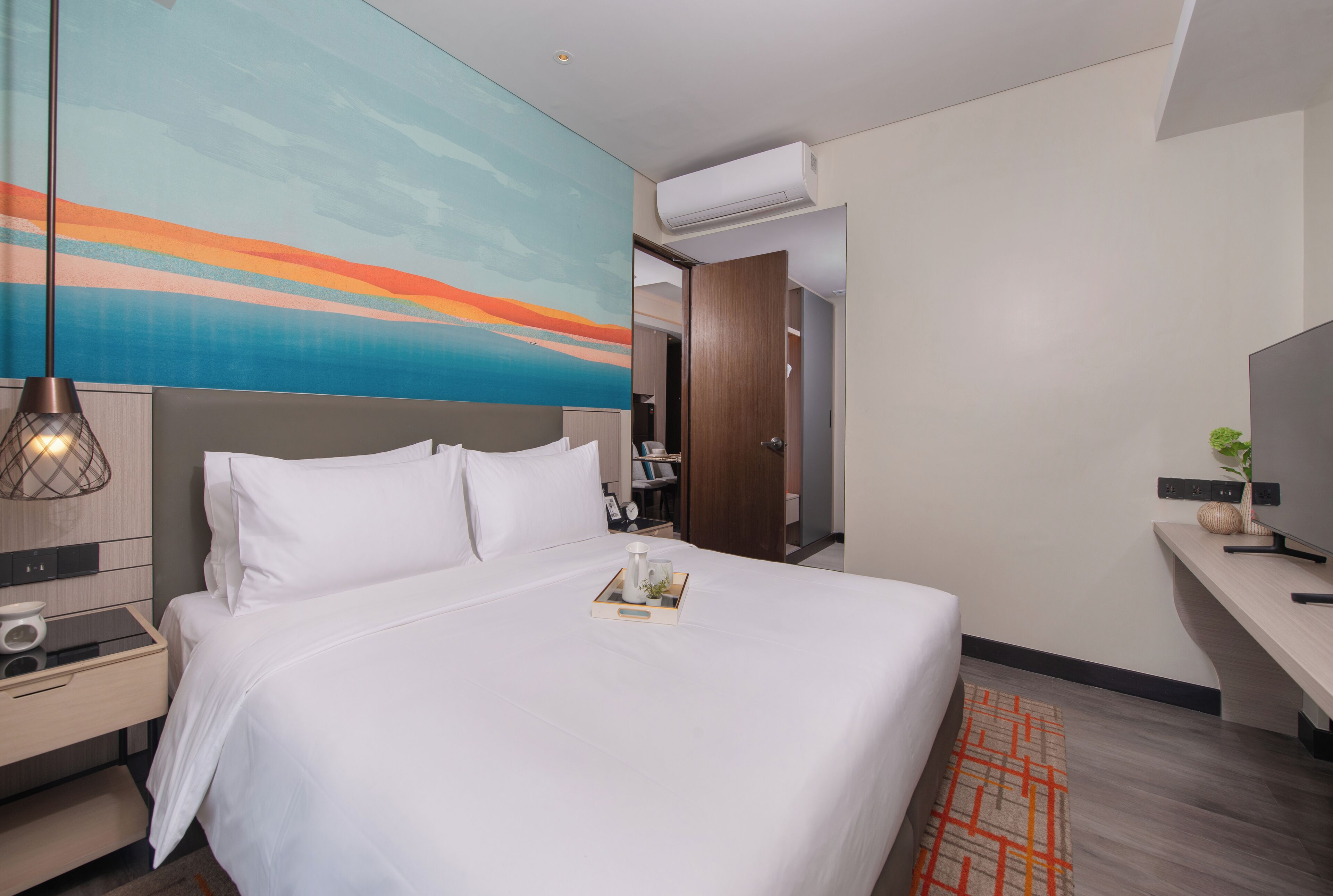 A modern hotel room with a large bed, a nightstand, a desk, and a painting on the wall.