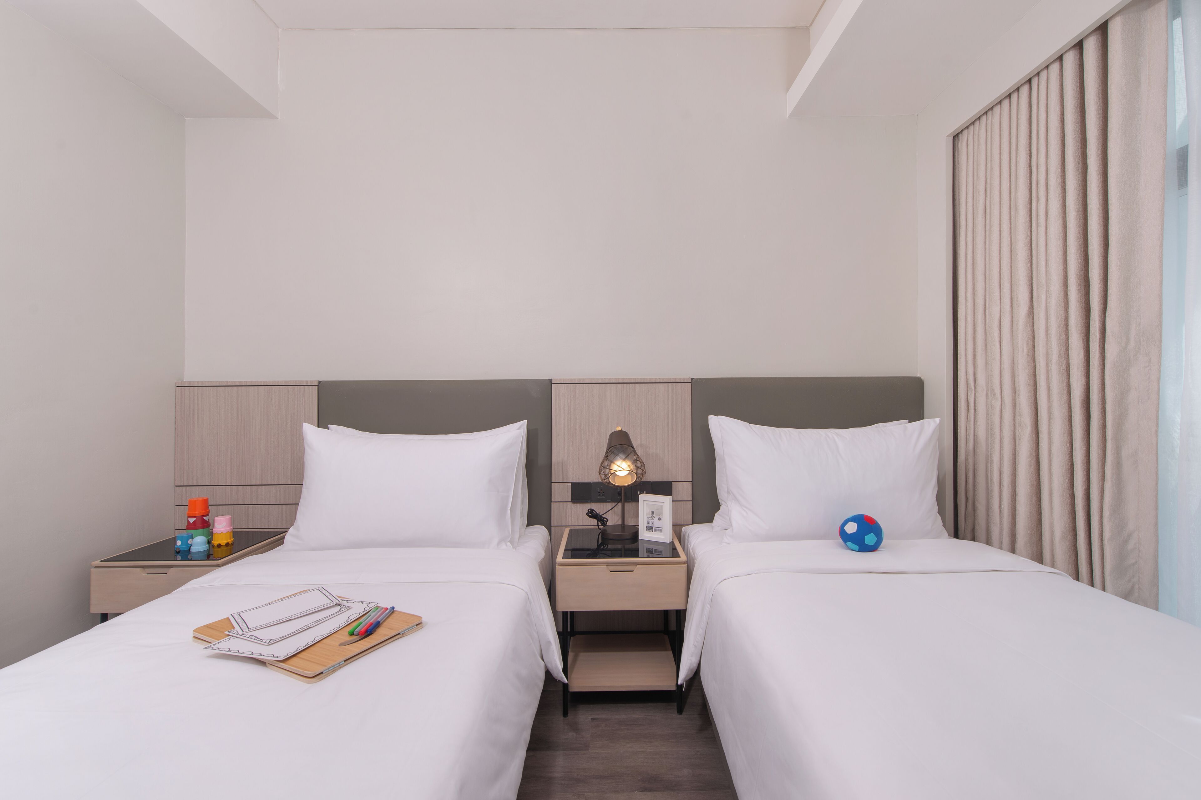 A hotel room with two single beds, a bedside table with a lamp, a nightstand with a lamp, and a window with curtains.