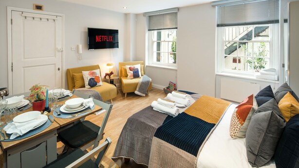 A well-lit room with a bed, a sofa, a TV mounted on the wall, and a dining area with a table set for two.