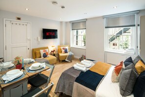 Een goed verlichte kamer met een bed, een bank, een aan de muur bevestigde televisie en een eethoek met een tafel voor twee personen.