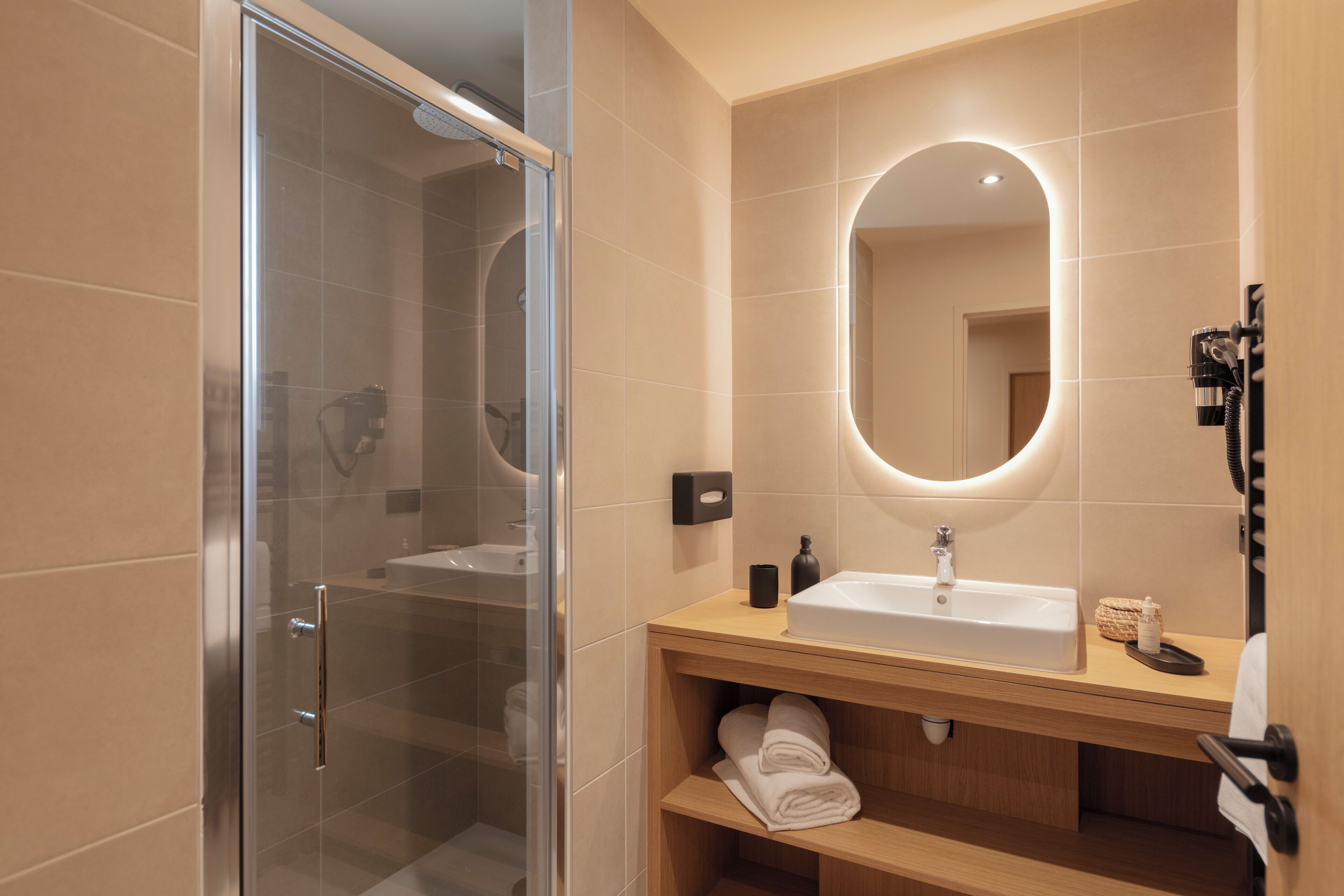 Un baño moderno con ducha envuelta en vidrio, tocador de madera con lavamanos y espejo con iluminación integrada.
