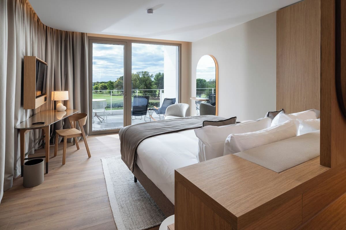 A modern hotel room with a large bed, a desk with a lamp, a TV, and a view of a balcony with outdoor seating.