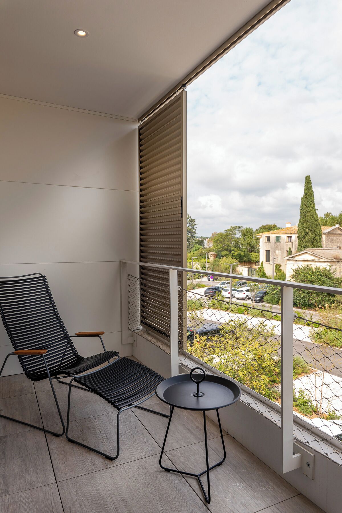 A balcony with a black metal chair and a small round table, overlooking a parking area and buildings.