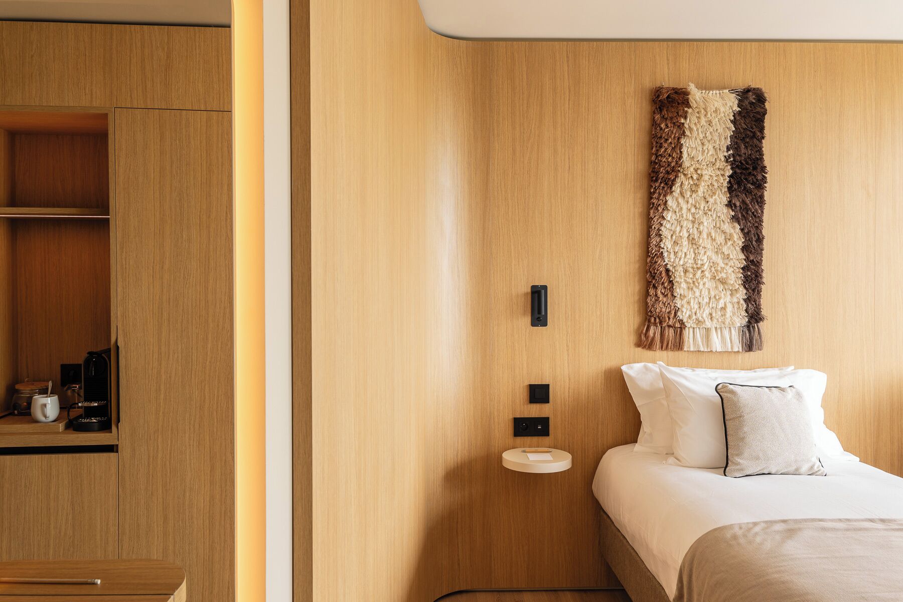 A modern hotel room with a wooden wardrobe, a bed with white bedding, and a textured wall hanging.