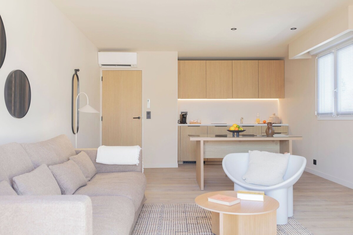 A modern living room with a sofa, a chair, a small table, and a kitchen area with wooden cabinets and a countertop.