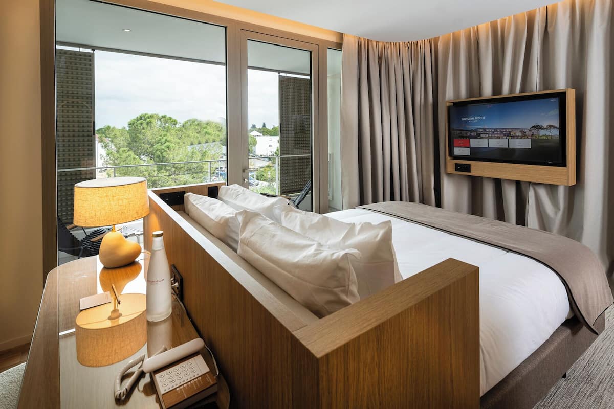 A hotel room with a bed, a TV, a desk with a lamp, and a window with a view of trees and buildings.
