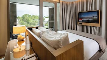 A hotel room with a bed, a TV, a desk with a lamp, and a window with a view of trees and buildings.