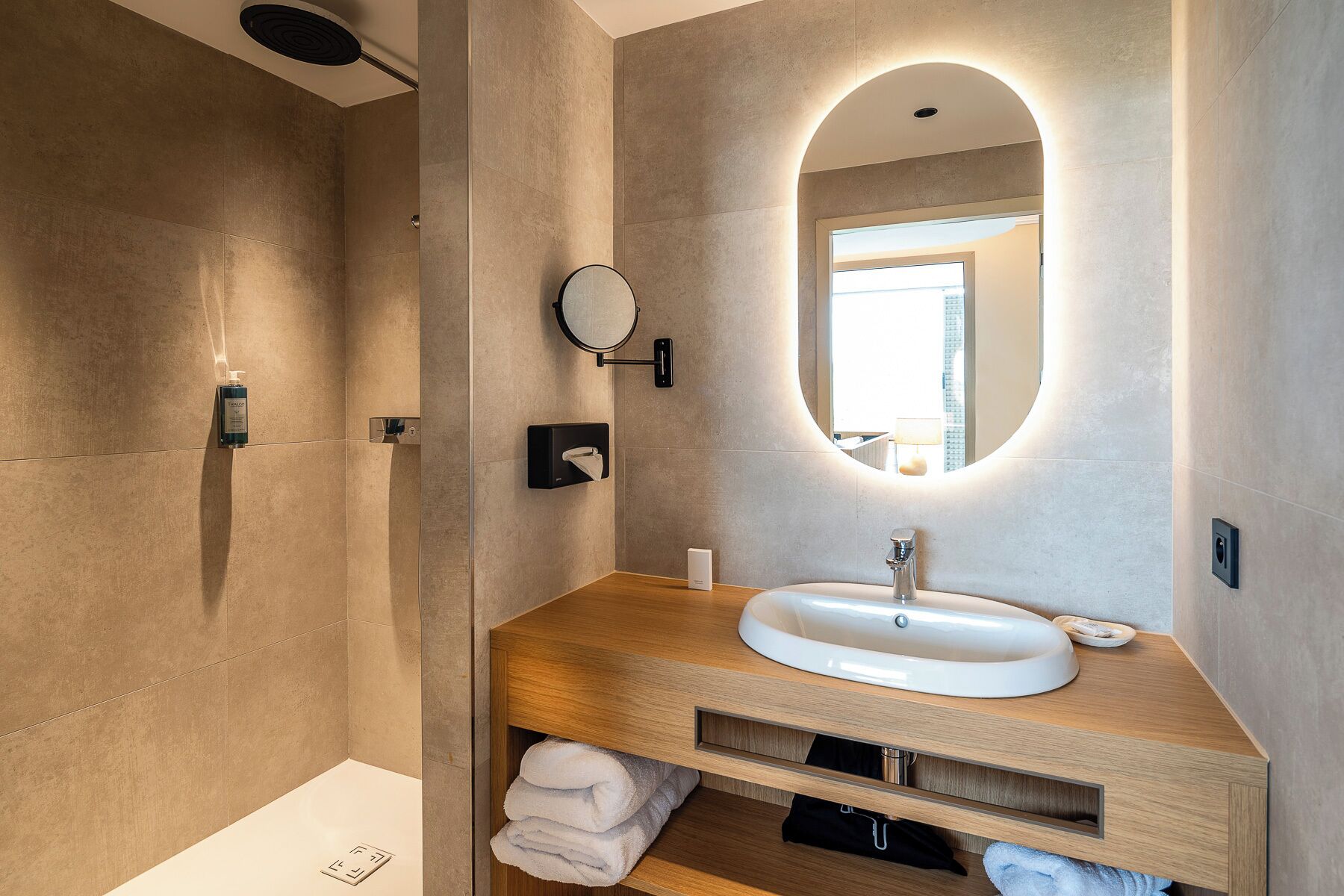 A modern bathroom with a large oval mirror, a white sink, and a shower area.