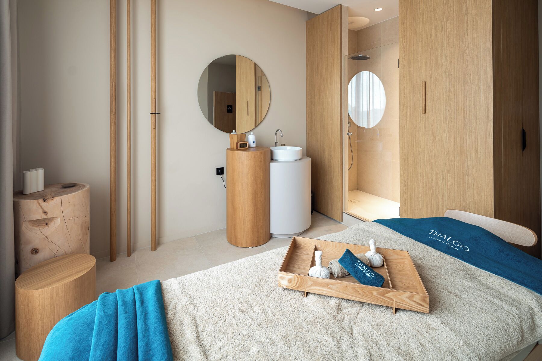 A modern hotel room with a wooden vanity, a round mirror, a bed with a blue towel, and a tray with toiletries.