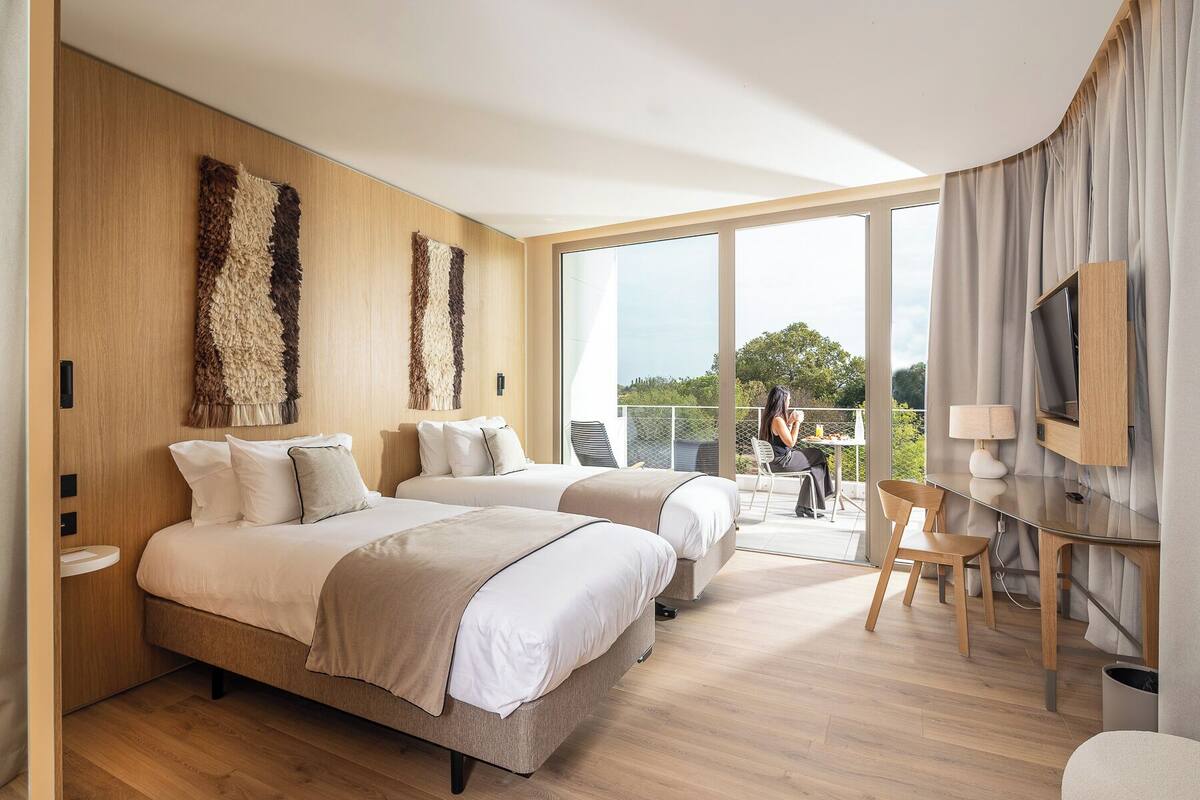 A modern hotel room with two beds, a balcony, a TV, and a dining area.