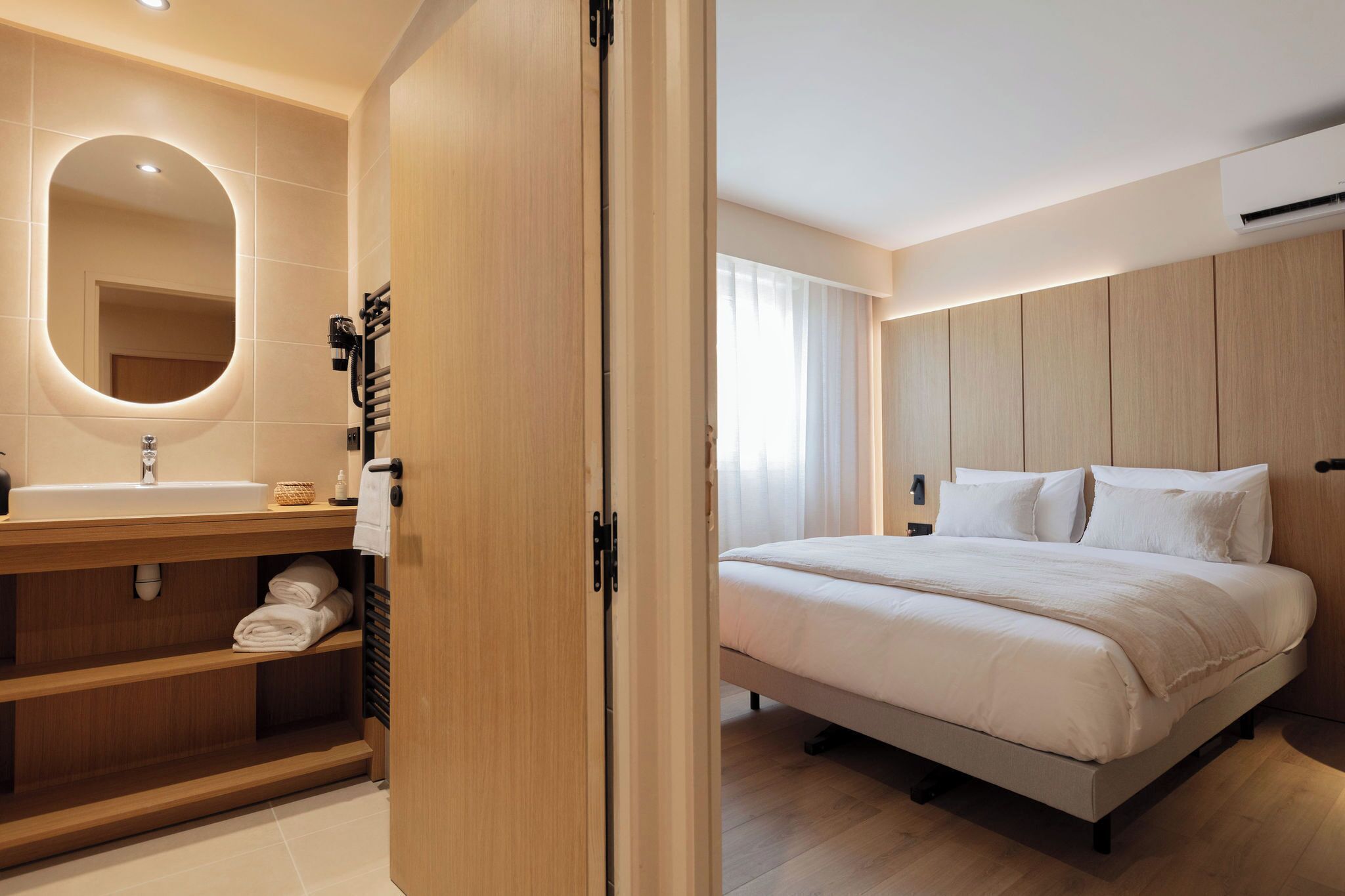 Ein modernes Hotelzimmer mit einem hölzernen Waschtisch, einem großen Spiegel, einem Bett mit weißer Bettwäsche und einem Holz-Kopfschild.