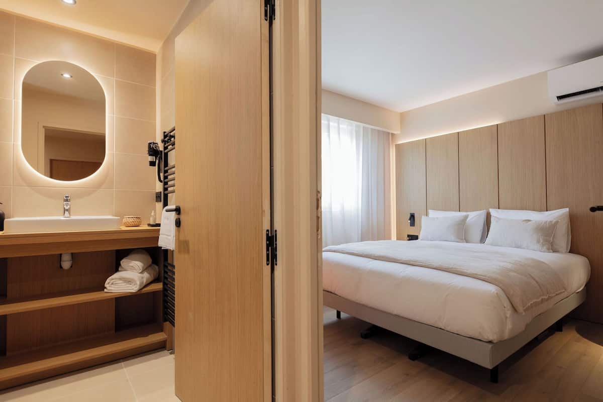 A modern hotel room with a wooden vanity, a large mirror, a bed with white bedding, and a wooden headboard.