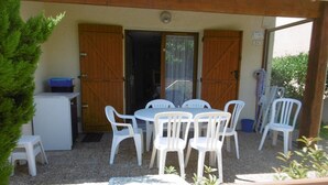Eine überdachte Terrasse mit einem runden Tisch und weißen Plastikstühlen, einem kleinen Kühlschrank und Holztüren.
