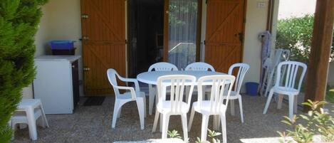 A covered outdoor patio with a round table and white plastic chairs, a small refrigerator, and wooden doors.