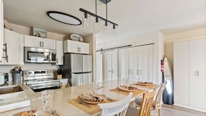 A modern kitchen with white cabinetry, stainless steel appliances, and a dining area with a long table set for four. - Studio with prime location next to Beach, Jolly Rogers and Boardwalk (Ocean City)