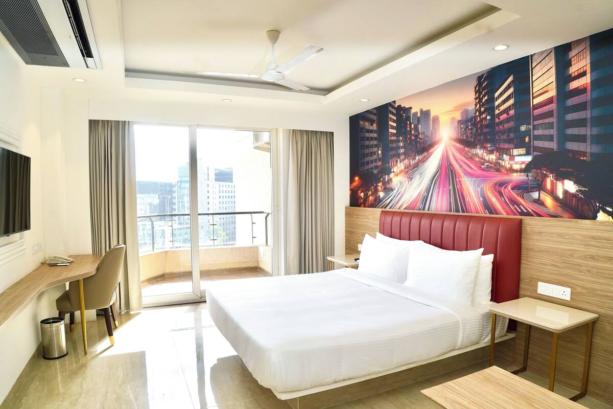 A hotel room with a large bed, a desk, a chair, a small table, and a view of the city through the window.