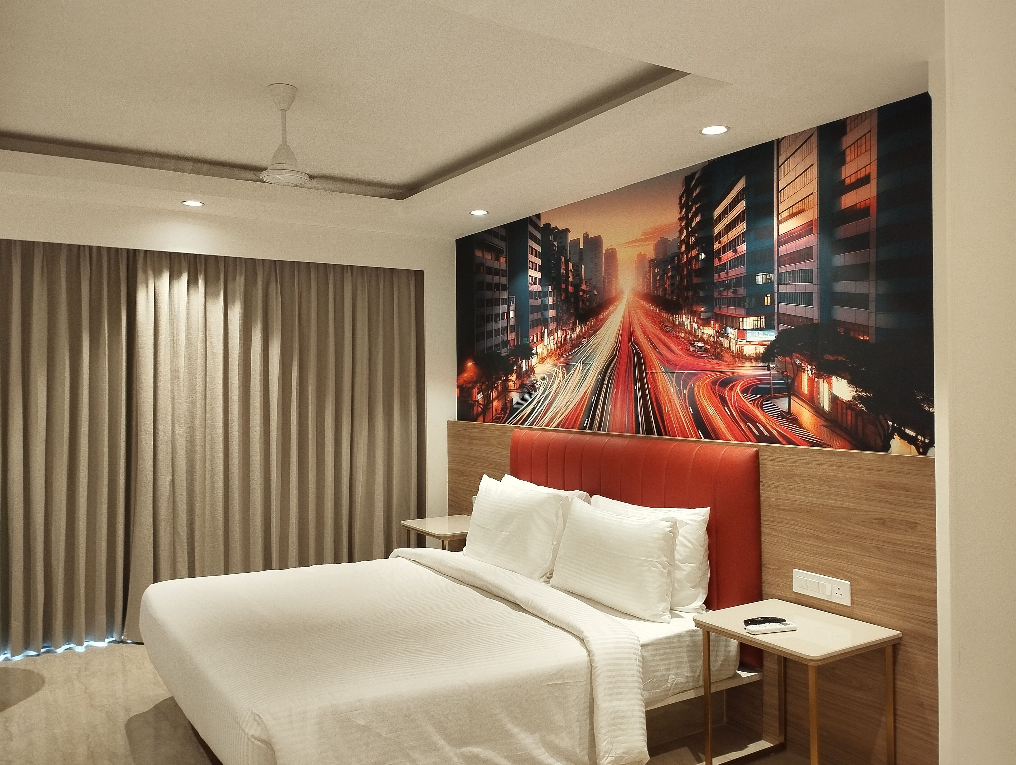 Una habitación de hotel moderna con una cama grande, un cabecero con un mural de un paisaje urbano, un ventilador de techo y una mesita de noche con un teléfono y un control remoto.