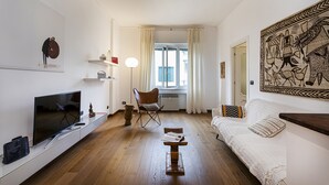 Ein Wohnzimmer mit Flachbildfernseher, einem Holztisch, einem weißen Sofa und einem Sessel. Ein Fenster mit Vorhängen, ein Heizkörper und ein Wandbild sind ebenfalls zu sehen.